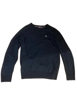 Mörkblå stickad tröja från Morris - Klassisk mörkblå stickad tröja från Morris i 100% merinoull. Tröjan har rund halsringning, ribbade muddar och en liten broderad logga på bröstet. Perfekt för lager på lager och enkel att matcha med olika stilar.