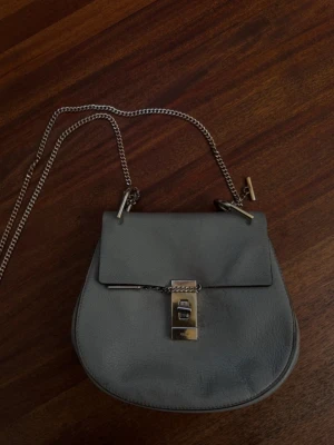  Chloé väska - Snygg grå axelväska från Chloé i mjukt läder med guldfärgad kedjerem och ikoniskt vridlås framtill. Väskan har rundad form och stilren design som passar till många outfits. Perfekt för dig som vill ha en trendig och lyxig accessoar.