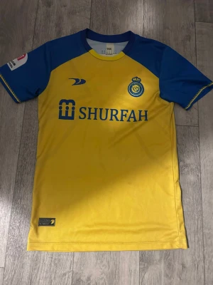 Al Nassr Ronaldo matchtröja gul/blå - Snygg gul och blå matchtröja från Al Nassr med Ronaldo och nummer 7 på ryggen. Tröjan har klubbmärke och sponsorlogga på bröstet samt blå detaljer på ärmar och axlar. Tillverkad i lätt och ventilerande material, perfekt för fotbollsträning eller match.
