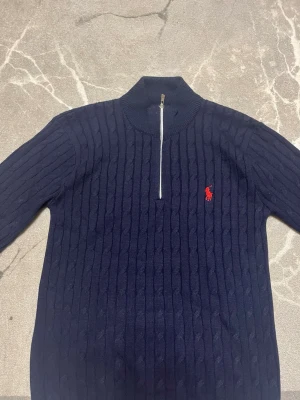 Mörkblå kabelstickad tröja Polo Ralph Lauren - Mörkblå kabelstickad tröja från Polo Ralph Lauren med halv dragkedja och röd broderad logga på bröstet. Tröjan har hög krage, långa ärmar och ribbade muddar. Perfekt för dig som gillar klassisk och stilren look. Står storlek L på tröjan men passar S.