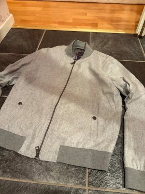 Grå bomberjacka från Tommy Hilfiger - Snygg grå bomberjacka från Tommy Hilfiger med ribbade muddar och krage. Jackan har dragkedja framtill, två fickor med knapp och diskret logga på insidan. Materialet är mjukt och har en lätt melerad look. Perfekt för dig som vill ha en stilren och modern jacka.