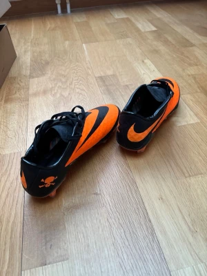 Nike Hypervenom orange fotbollsskor - Säljer ett par Nike Hypervenom fotbollsskor i svart och orange med klassisk Swoosh-logga och dödskalle-detalj på hälen. Skorna har snörning och är tillverkade i syntetmaterial för bra grepp och kontroll på planen. Perfekta för dig som vill sticka ut på matchen!