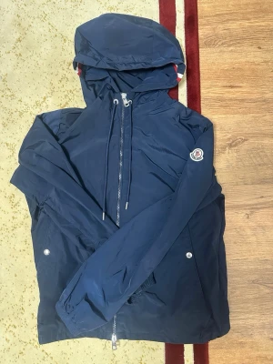 Mörklbå moncler windbreaker  - Snygg mörkblå vindjacka från Moncler med huva och dragsko. Jackan har silverfärgade detaljer, två sidofickor med tryckknappar och Moncler-logga på ärmen. Materialet är tunt och vattenavvisande, perfekt för blåsiga dagar.