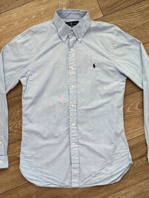 Ljusblå skjorta från Ralph Lauren - Klassisk ljusblå skjorta från Ralph Lauren med button-down krage och broderad logga på bröstet. Skjortan har långa ärmar, vita knappar och är tillverkad i mjuk bomull. Perfekt för en clean och stilren look. Oxford 