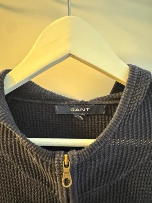 Mörkblå ziptröja från GANT - Mörkblå stickad ziptröja från GANT med grov struktur och en stor G-brodyr på bröstet. Tröjan har hel dragkedja framtill, ribbade muddar och en klassisk collegekänsla. Perfekt för dig som gillar sportig och avslappnad stil.