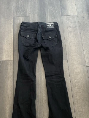 True Religion jeans 24 - Säljer ett par svarta jeans från True Religion med bootcut passform. Jeansen har två bakfickor med lock och knapp, samt klassiska framfickor. Snygg detalj med logopatch bak i midjan. Perfekta för en trendig och avslappnad stil.