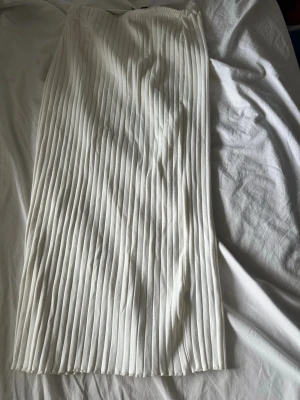 Vit plisserad långkjol - Säljer en elegant vit långkjol med plisserade veck. Kjolen har en rak och stilren siluett som ger en modern look. Perfekt att matcha med en crop top eller stickad tröja för en trendig outfit.