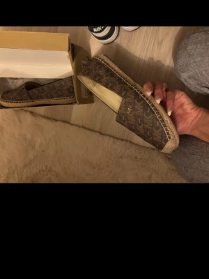 Bruna ballerinas från Michael Kors - Snygga bruna ballerinas från Michael Kors med klassiskt MK-mönster över hela skon. Skorna har en rund tå och dekorativt flätad kant i beige, vilket ger en trendig look. Materialet är canvas med detaljer i jute. Perfekt för dig som gillar stilrena och moderna skor.