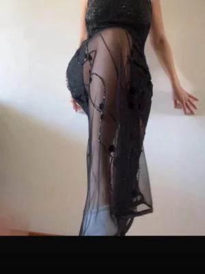 Svart glittrig långklänning med mesh - Svart långklänning med glittrigt tyg och transparent mesh över ben och bröst. Klänningen har blommiga detaljer på meshpartiet och en figurnära passform. Perfekt för dig som vill sticka ut med en elegant och unik look.