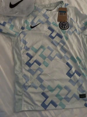 Inter Milan matchtröja Nike ljusblå - Snygg Inter Milan matchtröja från Nike i ljusblått med geometriskt mönster i blå och turkosa nyanser. Tröjan har korta ärmar, rund hals och klubbmärke på bröstet. Perfekt för dig som älskar fotboll och vill ha en unik look på planen.
