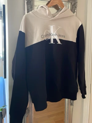 Svart och beige hoodie från Calvin Klein i nyskick!! - Snygg hoodie från Calvin Klein Jeans i svart och beige med stor CK-logga på bröstet. Hoodien har en klassisk huva och är gjord i mjukt bomullsmaterial. Perfekt för dig som gillar stilrena och trendiga plagg med tydlig logotyp.