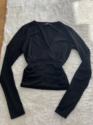 Svart långärmad topp med djup v-ringning - Säljer en svart långärmad topp med v-ringning och figurnära passform. Materialet är mjukt och stretchigt, perfekt för att skapa en snygg siluett. Enkel och stilren design som är lätt att matcha med olika outfits.finns ett väldigt litet hål vid lappen som knappt syns (se bild). 