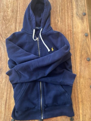 Mörkblå hoodie från Polo Ralph Lauren - Säljer en mörkblå hoodie med dragkedja från Polo Ralph Lauren. Hoodien har huva med vita snören, ribbade muddar och fickor framtill. Materialet är mjukt och bekvämt, perfekt för en avslappnad look. Ikoniska Polo-loggan i gult på bröstet.