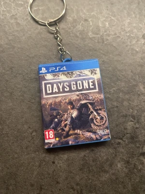 Days Gone nyckelring PS4 - Cool nyckelring med motiv från PS4-spelet Days Gone. Nyckelringen är formad som ett miniatyrspelcase i blå plast med detaljerad framsida och baksida. Perfekt för gamers som vill visa sitt intresse för spel och PlayStation.