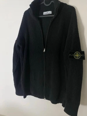 Svart stickad ziptröja Stone Island - Stickad svart ziptröja från Stone Island med ribbad struktur och hög krage. Tröjan har dragkedja framtill och den ikoniska Stone Island-loggan på ärmen. Tröjan är icke äkta!