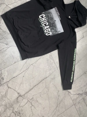 Svart hoodie med Chicago-tryck - Cool svart hoodie med stor Chicago-tryck på bröstet och texten 'Always Looking Forward' på ena ärmen. Klassisk huva och magficka framtill. Materialet känns mjukt och bekvämt, perfekt för chill dagar. Snygg urban stil med grafiskt motiv.