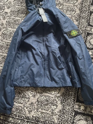 Blå Stone Island vindjacka med huva - Snygg blå vindjacka från Stone Island med huva och dragkedja framtill. Jackan har det klassiska Stone Island-märket på vänster ärm och är tillverkad i ett lätt och slitstarkt material. Perfekt för att hålla vinden borta under kyliga dagar.