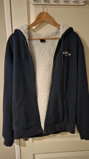 Mörkblå hoodie från Blakely - Mysig mörkblå hoodie från Blakely med vit teddyfoder på insidan. Hoodien har dragkedja, två fickor och en huva med snören. Märket är broderat på bröstet och materialet är mjukt och skönt.
