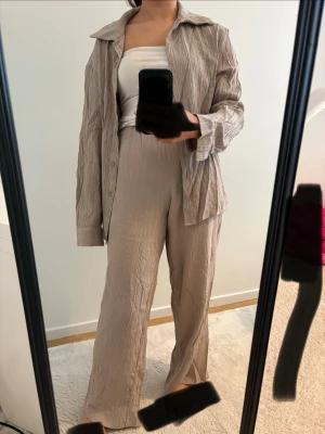 Beige loungewear-set med skjorta - Säljer ett beige loungewear-set med matchande skjorta och vida byxor i krinklat material. Skjortan har klassisk krage och kan bäras både öppen och knuten. Perfekt för chill dagar hemma. Materialet är mjukt och luftigt.