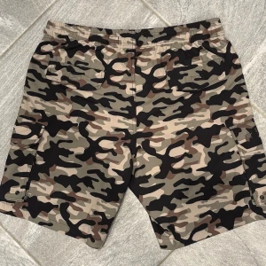 Kamouflagemönstrade badbyxor - Snygga och praktiska badbyxor med nätfoder  - Midjeband med dragsko - Två framfickor - Två påsydda sidofickor - höger ficka med logo och dragkedja. - Två bakfickor   Benläng från resår: 47 cm Innerbenslängd: 20,5 cm Benvidd 33 cm   