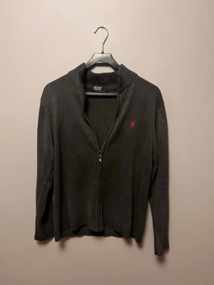 Mörkgrå zip-kofta från Polo Ralph Lauren - Mörkgrå stickad kofta med dragkedja från Polo Ralph Lauren. Klassisk design med ribbade muddar och röd broderad logotyp på bröstet. Perfekt för lager-på-lager och snygg till jeans. Materialet är mjukt och bekvämt.