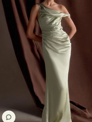 Grön oneshoulder långklänning - Elegant långklänning i pastellgrön satin med oneshoulder-design och draperade detaljer vid midjan. Klänningen har en slim passform och en lätt glansig yta som ger en lyxig känsla. Perfekt för bal eller festliga tillfällen.