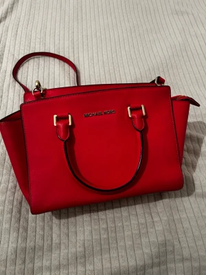 Röd handväska från Michael Kors - Snygg röd handväska från Michael Kors med guldfärgade detaljer och både handtag och axelrem. Väskan är tillverkad i skinn och har dragkedja upptill samt flera fack inuti för smart förvaring. Perfekt storlek för allt du behöver ha med dig.