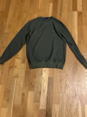 Olivgrön sweatshirt från Tommy Hilfiger - Olivgrön sweatshirt från Tommy Hilfiger med rund halsringning och diskret logga broderad på bröstet. Tröjan har ribbade muddar vid ärmslut och nederkant. Perfekt för en avslappnad och stilren look.