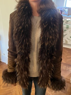 Brun pälsjacka med fluffig krage - Säljer en brun vintage pälsjacka med superfluffig, långhårig fuskpäls längs kragen och ärmsluten. Använda fåtal gånger så är i mycket gott skick😊