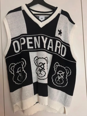 Svartvit stickad väst från OY - Cool stickad väst från OY med svartvita block och stora björnmotiv framtill. Västen har v-ringad hals och texten OPENYARD över bröstet. Perfekt för att skapa en unik och trendig look. Köpt i Tokyo.