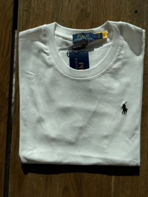 Vit t-shirt från Polo Ralph Lauren - Klassisk vit t-shirt från Polo Ralph Lauren med rund halsringning och den ikoniska svarta logobroderingen på bröstet. Tillverkad i mjuk bomull och har en normal passform. Perfekt basplagg för en clean och stilren look.