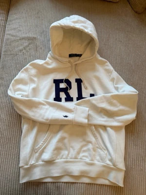 Vit hoodie från Polo Ralph Lauren - Vit hoodie från Polo Ralph Lauren med stor RL-logga i mörkblått på bröstet och klassisk broderad polospelare på ärmen. Hoodien har huva med dragsko, ribbade muddar och känguruficka. Materialet är mjukt och bekvämt bomullstyg.