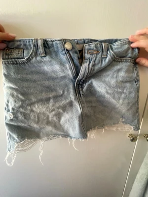 Ljusblå jeanskjol med fransig kant - Säljer en ljusblå jeanskjol med rå, fransig kant och slitna detaljer. Kjolen har klassisk femficksmodell, knapp- och dragkedjestängning samt bälteshällor. Perfekt för en avslappnad och trendig look.