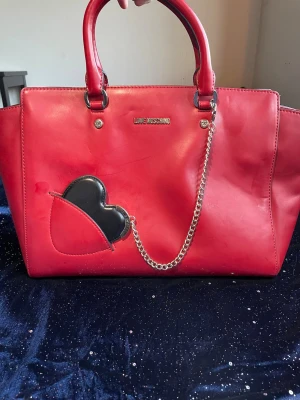 Röd handväska från Love Moschino - Cool röd handväska från Love Moschino i slätt konstläder. Väskan har två handtag, silverfärgade detaljer och en dekorativ kedja. På framsidan finns ett svart hjärta i en röd ficka som ger en lekfull touch. Perfekt för dig som vill sticka ut!