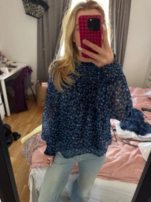 Blommig blå transparent blus - Säljer en blå blus med blommigt mönster i olika blå nyanser. Blusen är transparent, har hög hals och långa ärmar med lätt puff. Materialet känns lätt och luftigt, perfekt för att styla med jeans eller kjol. Märke ej synligt.