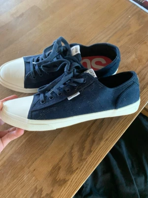 Mörkblå Superdry canvas sneakers - Snygga mörkblå sneakers från Superdry med klassisk låg profil. Skorna har vita gummisulor och tåhättor samt matchande mörkblå skosnören. Tillverkade i slitstark canvas med Superdry-logga på sidan och plös. Perfekta för en avslappnad streetstil.