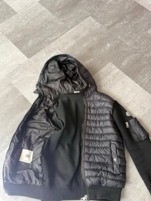Svart dunjacka med huva från Moncler - Säljer en svart dunjacka från Moncler med quiltad framsida och stickade ärmar. Jackan har en huva, dragkedja framtill och Moncler-logga på ärmen. Materialet är en kombination av dun och stickad textil, vilket ger både värme och stil.                      Små slitage finns, skickar bild vid intresse.        Priset är inte fast 😃