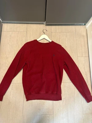  tröja från  Ralph Lauren - Snygg mörkröd tröja från Polo Ralph Lauren med klassisk rund halsringning och diskret blå logga broderad på bröstet. Tröjan har långa ärmar och ribbade muddar vid hals, ärmslut och nederkant. Perfekt för dig som gillar stilrena plagg.
