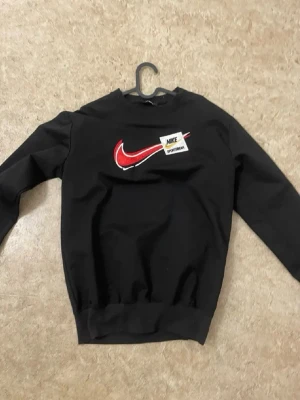 Svart sweatshirt från Nike - Säljer en svart sweatshirt från Nike med röd och vit swoosh-logga och en patch med texten 'Nike Sportswear' på bröstet. Tröjan har rund hals, långa ärmar och ribbade muddar. Perfekt för en sportig och avslappnad stil.