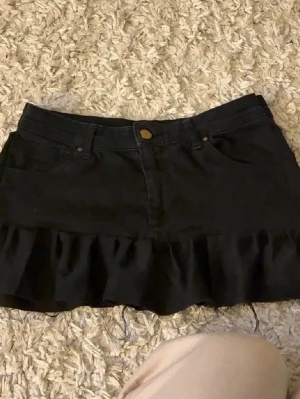 Svart jeanskjol med volangkant - Säljer en svart jeanskjol med volangkant nertill. Kjolen har klassisk femficksdesign, knapp och dragkedja framtill. Materialet är denim och modellen är kort och figurnära med en söt volangdetalj längst ner.