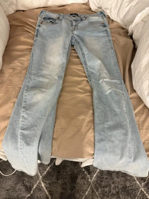 Ljusblå bootcut jeans True Religion - Säljer ett par ljusblå bootcut jeans från True Religion med ikoniska broderade fickor bak i orange och vitt. Jeansen har klassisk femficksmodell, låg midja och är tillverkade i denim med lätt tvättad look. Perfekta för dig som gillar retrovibbar och streetstyle.