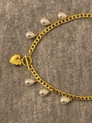 Guldfärgat halsband med pärlor och hjärta - Snyggt guldfärgat halsband med droppformade vita pärlor och ett hjärtformat hänge. Kedjan är chunky och har ett stilrent toggle-lås. Perfekt för dig som vill ha något unikt och trendigt. Materialet är rostfritt stål och pärlor.
