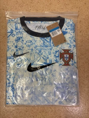 Portugal Nike fotbollströja blå/vit - Säljer en Portugal fotbollströja från Nike i blått och vitt med grafiskt mönster. Tröjan har svarta detaljer vid krage och ärmslut samt Portugals landslagsemblem på bröstet. Perfekt för dig som älskar fotboll och vill sticka ut på planen.