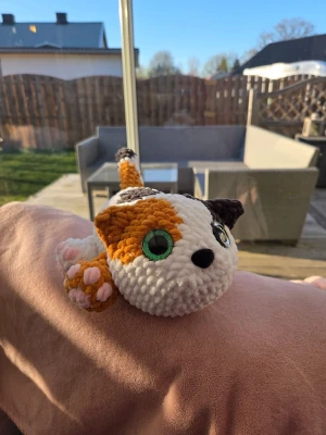 Virkad katt med stora gröna ögon - Handgjord virkad katt i vitt, orange och brunt garn. Katten har stora glittriga gröna säkerhetsögon, rosa nos och tassar samt en lång svans. Mjuk och gosig, perfekt som dekoration eller gosedjur. Unik och charmig design som sticker ut.