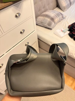 Grå väska Lacoste  - Snygg grå väska från Lacoste i stilren design med mjukt konstlädermaterial. Väskan har breda axelband och praktiskt innerfack med mesh-detaljer. Perfekt för dig som gillar en clean och modern look.