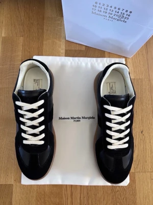 Maison Margiela svarta sneakers - Svarta sneakers från Maison Martin Margiela med vita snören och beige innersula. Skorna har en klassisk låg silhuett, ovandel i mocka och läder samt en brun gummisula. Perfekta för dig som gillar stilrena och tidlösa sneakers. Om du har funderingar e det ba o höra av dig