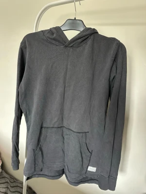 Svart hoodie med känguruficka - En enkel svart hoodie med huva och känguruficka framtill. Tröjan har långa ärmar och är tillverkad i ett mjukt bomullsmaterial. Perfekt för dig som gillar en avslappnad och stilren look. Liten etikett nertill på framsidan.