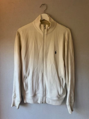 Beige ziptröja från Polo Ralph Lauren - Snygg beige ziptröja från Polo Ralph Lauren med hög krage och diskret logga broderad på bröstet. Tröjan har två fickor framtill och är tillverkad i mjukt material. Perfekt för lager-på-lager och chill stil.