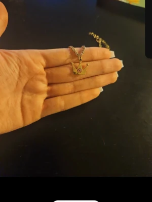 Guldigt halsband med krona och rosa sten - Säljer ett guldigt halsband med ett hängsmycke i form av en krona. Kronan har en liten rosa sten i mitten som ger en extra touch. Kedjan är tunn och elegant, perfekt för dig som gillar unika detaljer. Materialet ser ut att vara metall.