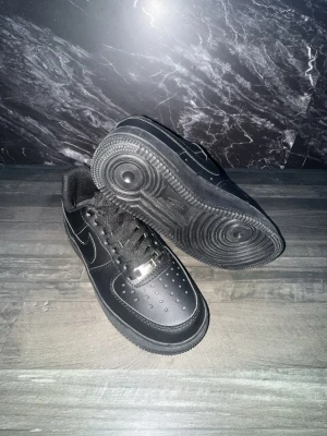 Nike Air Force 1 Low helsvart läder - Klassiska Nike Air Force 1 Low sneakers i helsvart läder med svart sula och svarta skosnören. Ikonisk Nike Swoosh på sidan och metallbricka med 'AF1' på snörningen. Perforerad tå för ventilation och tidlös streetwear-look.